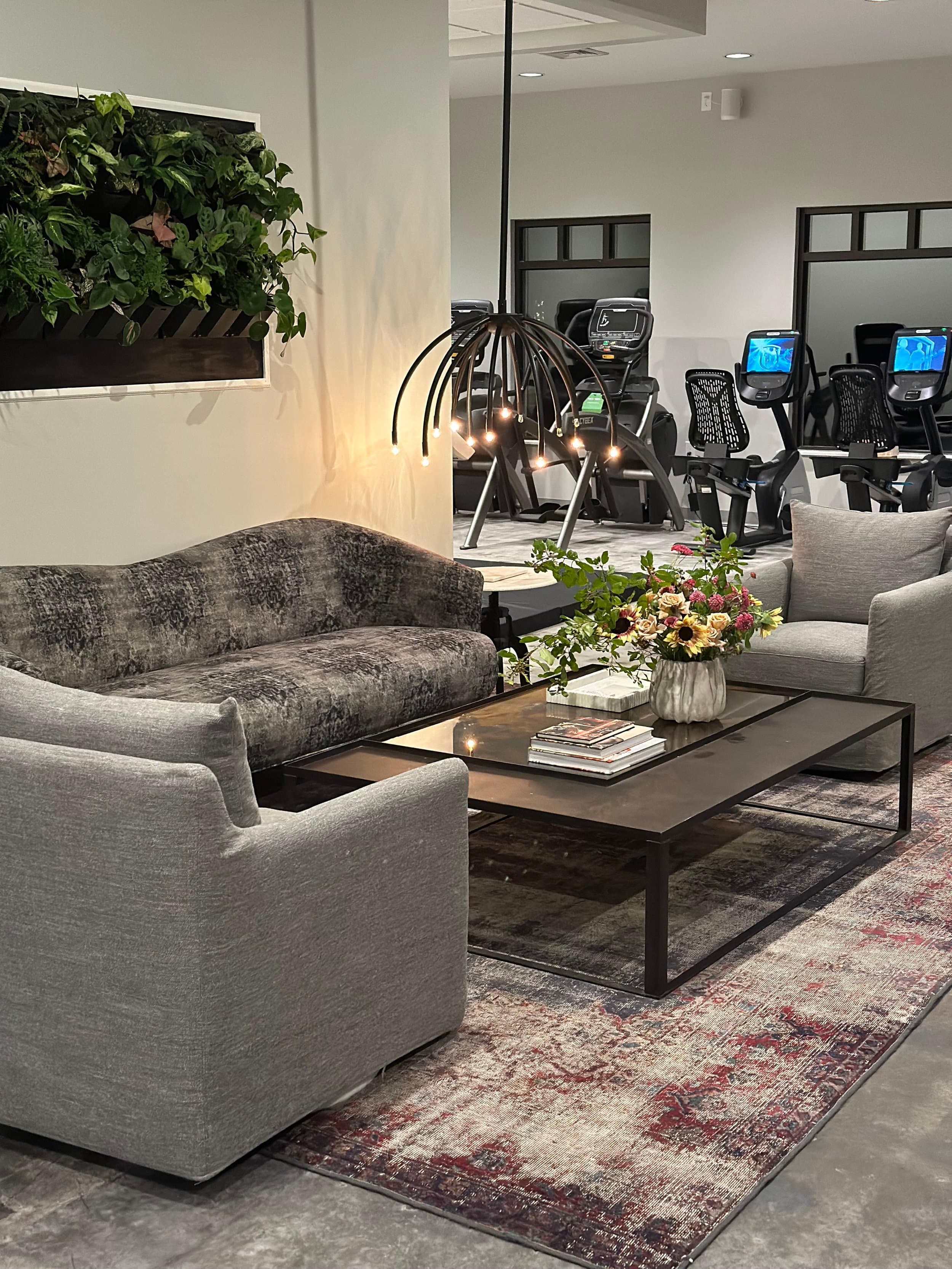 The members' lounge at LX Fitness Club — upholstered seating and a living plant wall, with the cardio floor visible through a glass partition beyond.