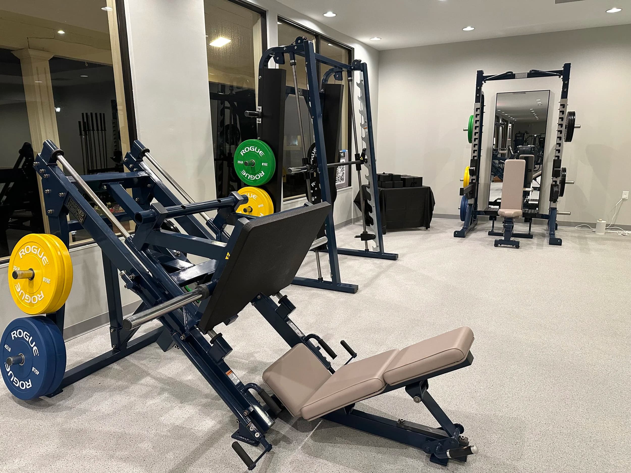 The strength area at LX Fitness Club — a Hammer Strength plate-loaded leg press, Rogue bumper plates, a half rack, and an adjustable bench.